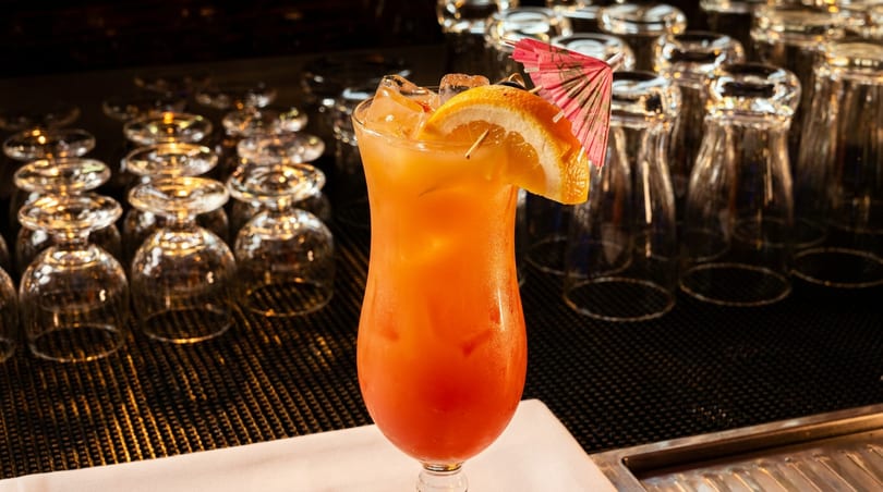 Tropical orange and red cocktail with an orange slice and pink umbrella on a bar.