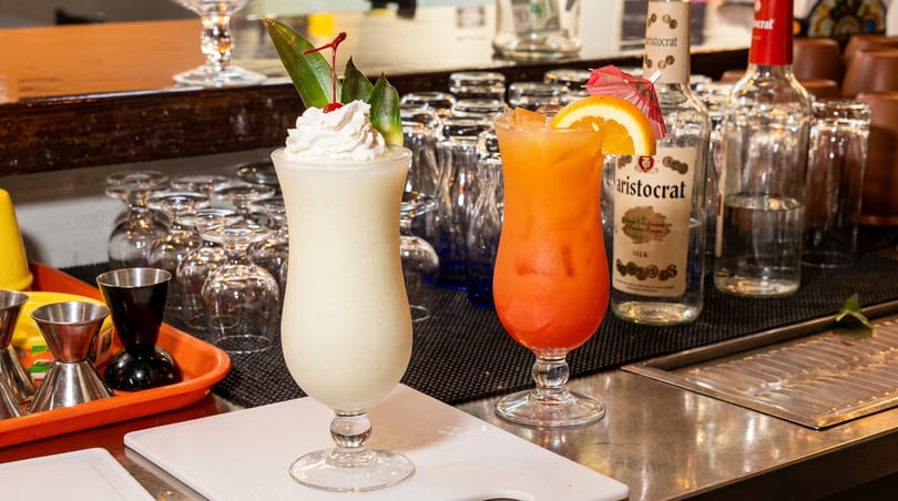 Two tropical cocktails, a white piña colada and an orange sunrise, garnished on a bar.