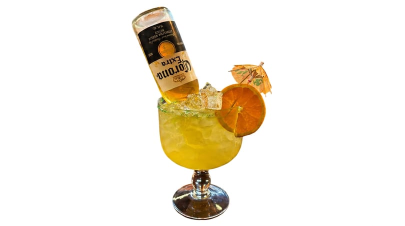 Corona Extra bottle inverted into a large, iced margarita with a green rim, orange slice, and umbrella.