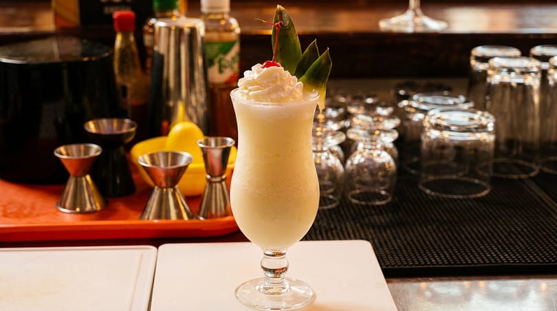 A blended piña colada cocktail with whipped cream, cherry, and pineapple leaves at a bar.