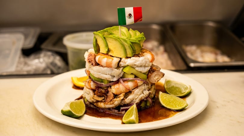 Stacked Mexican seafood tower with shrimp, avocado, lime, and a small flag on a white plate.