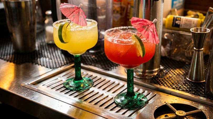 Yellow and red margaritas with lime wedges and umbrellas on a bar counter.