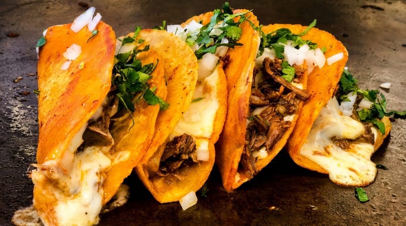 Four cheesy birria tacos with shredded meat, onions, and cilantro on a dark surface.