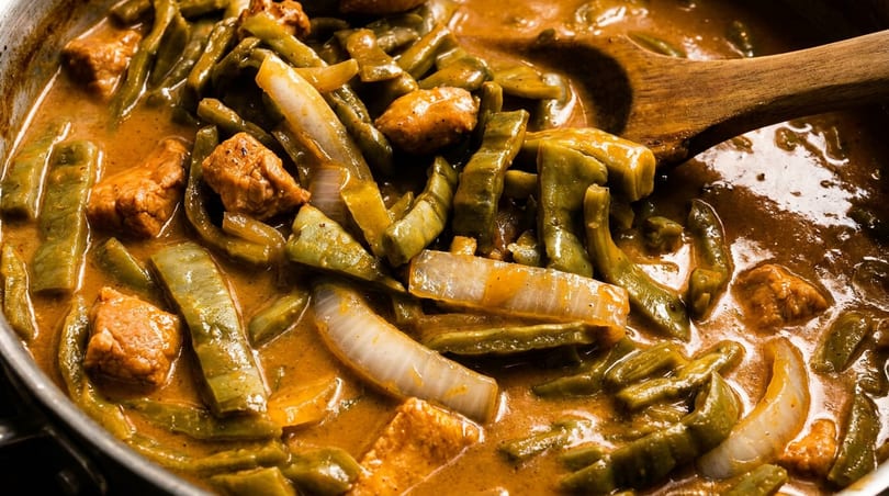 Nopales and pork stew with onions in a rich red sauce, stirred by a wooden spoon.